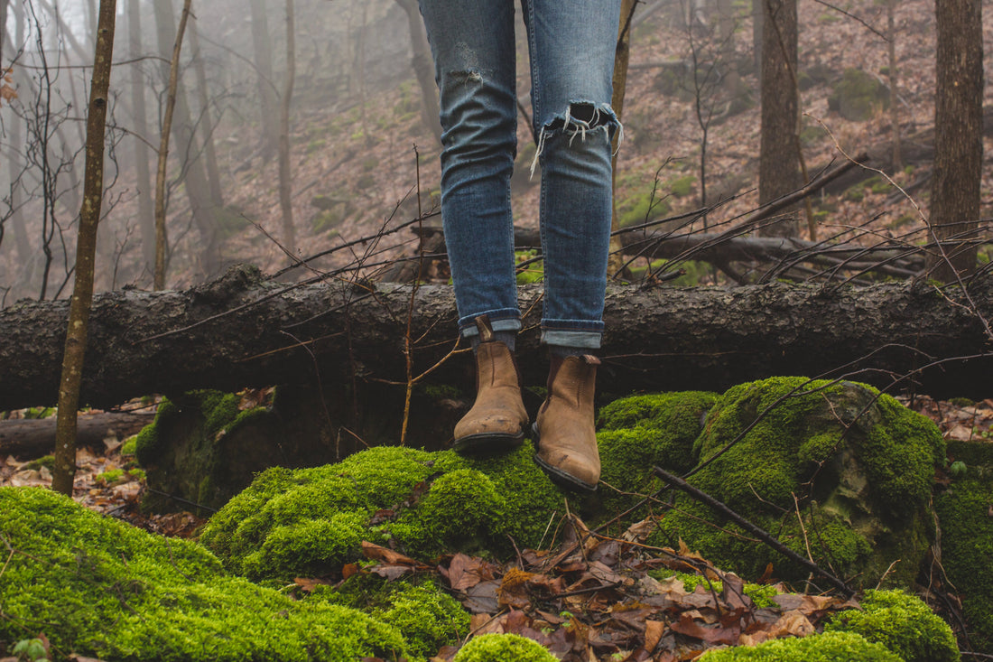 Person wearing jeans and boots standing on a mossy rock in the woods